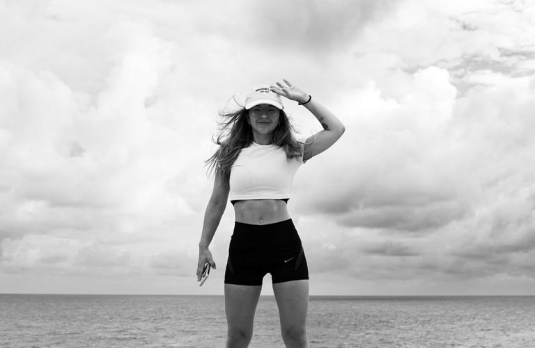 Woman in sportswear stands confidently by the sea against a cloudy sky.