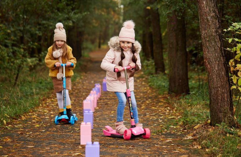 Two girls having fun on scooters in a leafy autumn park trail.